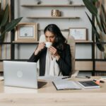 Woman sipping coffee while working on a laptop in a modern office.