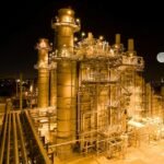 An industrial power plant illuminated at night with a full moon in the sky