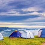 Plusieurs tentes de camping installées sur herbe verte avec montagnes et nuages en arrière-plan au lever du soleil